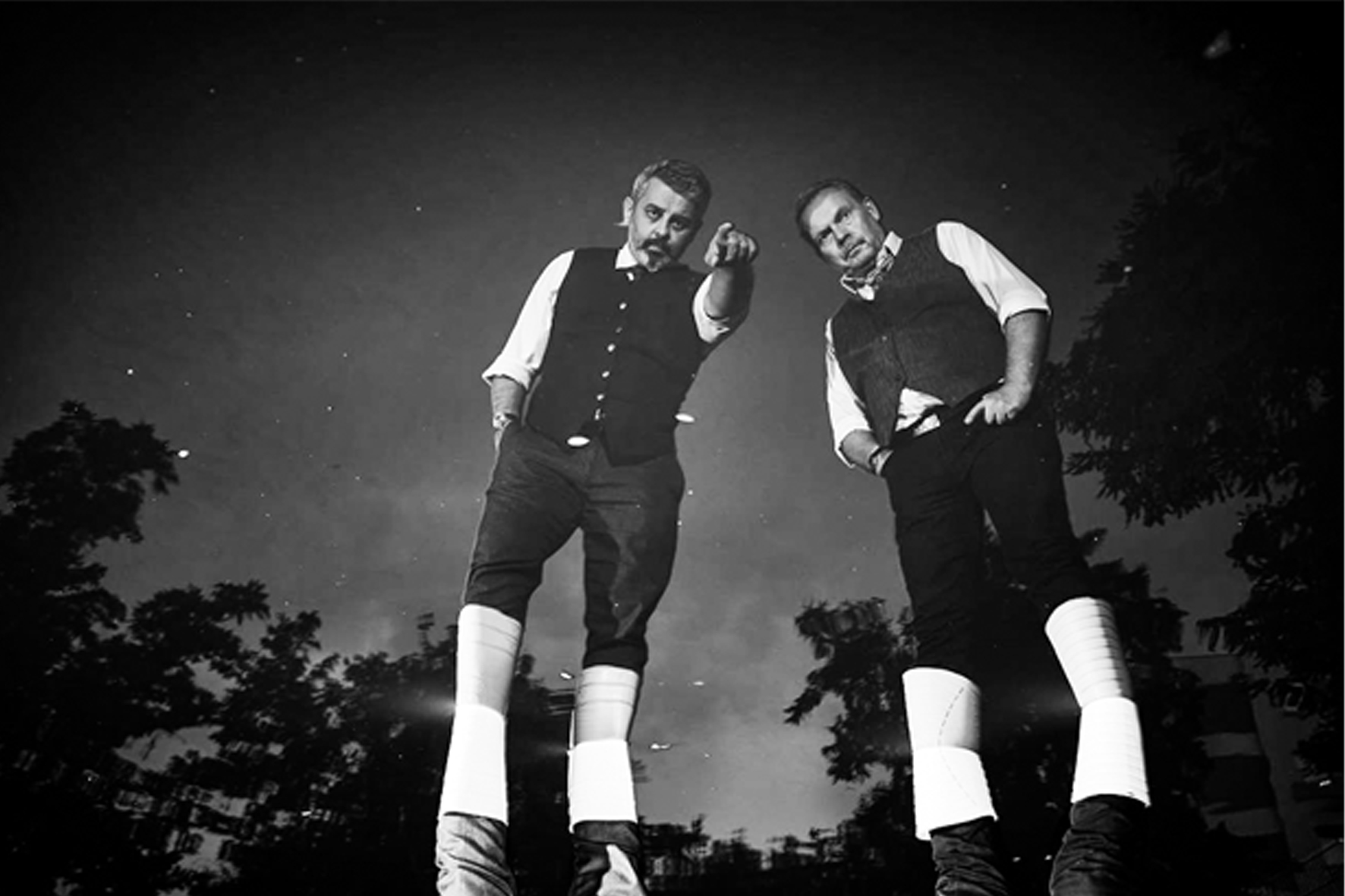 zdjęcie czarno-białe; dwóch mężczyzn w białych podkolanówkach, czarnych spodniach do kolan, białych koszulach i czarnych kamizelkach stoją na tle koron drzew. Jeden z nich wskazuje przed siebie palcem